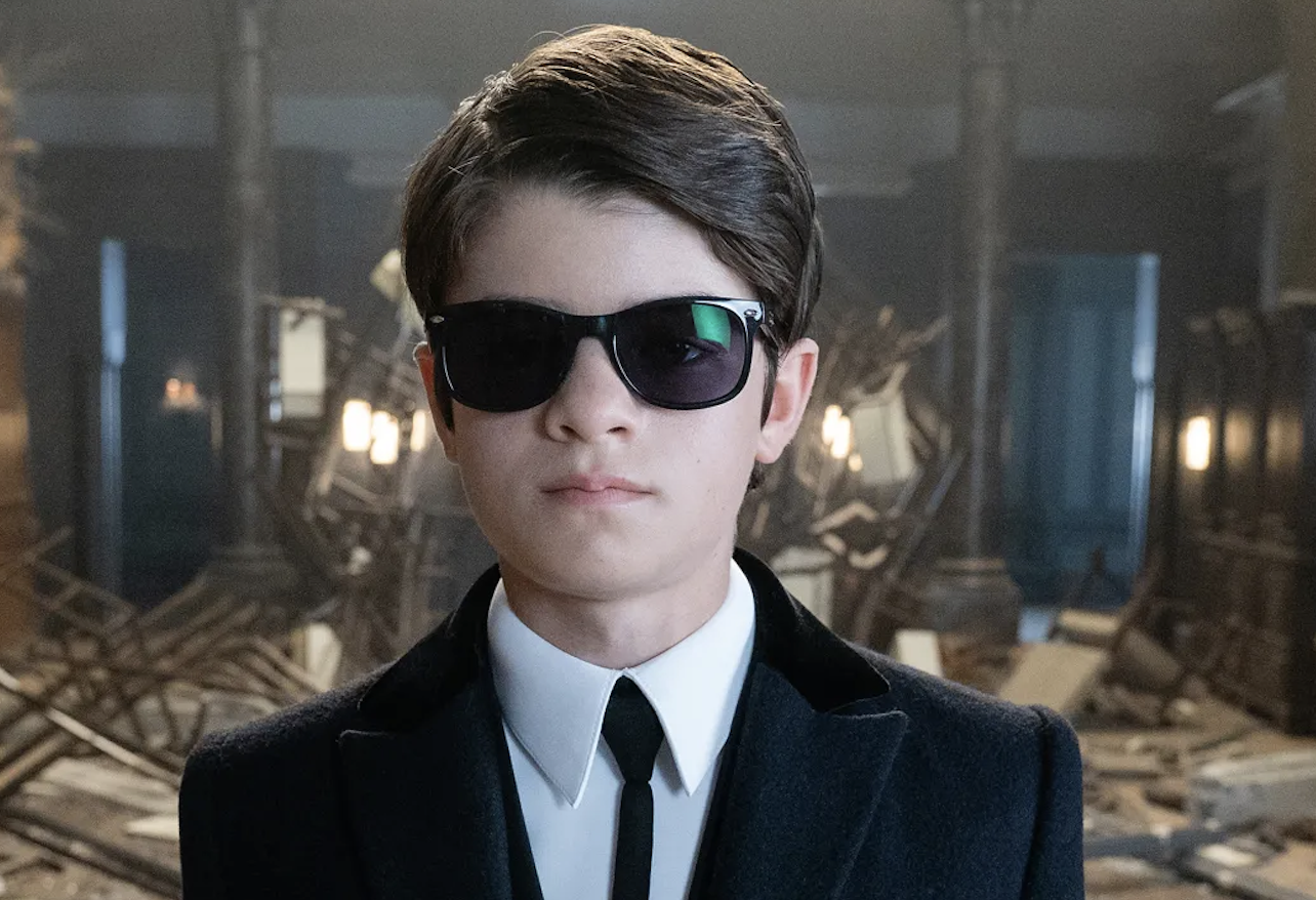 Person wearing dark sunglasses and a formal suit with a tie, standing in a dimly lit room with debris and a mysterious atmosphere