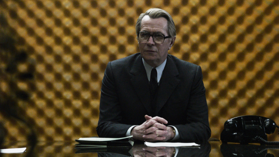 Gary Oldman is sitting at a desk, wearing a suit and glasses, with his hands clasped. Background shows a padded wall pattern