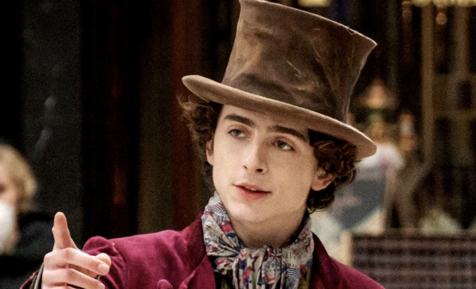 Timothée Chalamet dressed as Willy Wonka, wearing a top hat, a colorful scarf, and a velvet coat, in a scene from the movie "Wonka"
