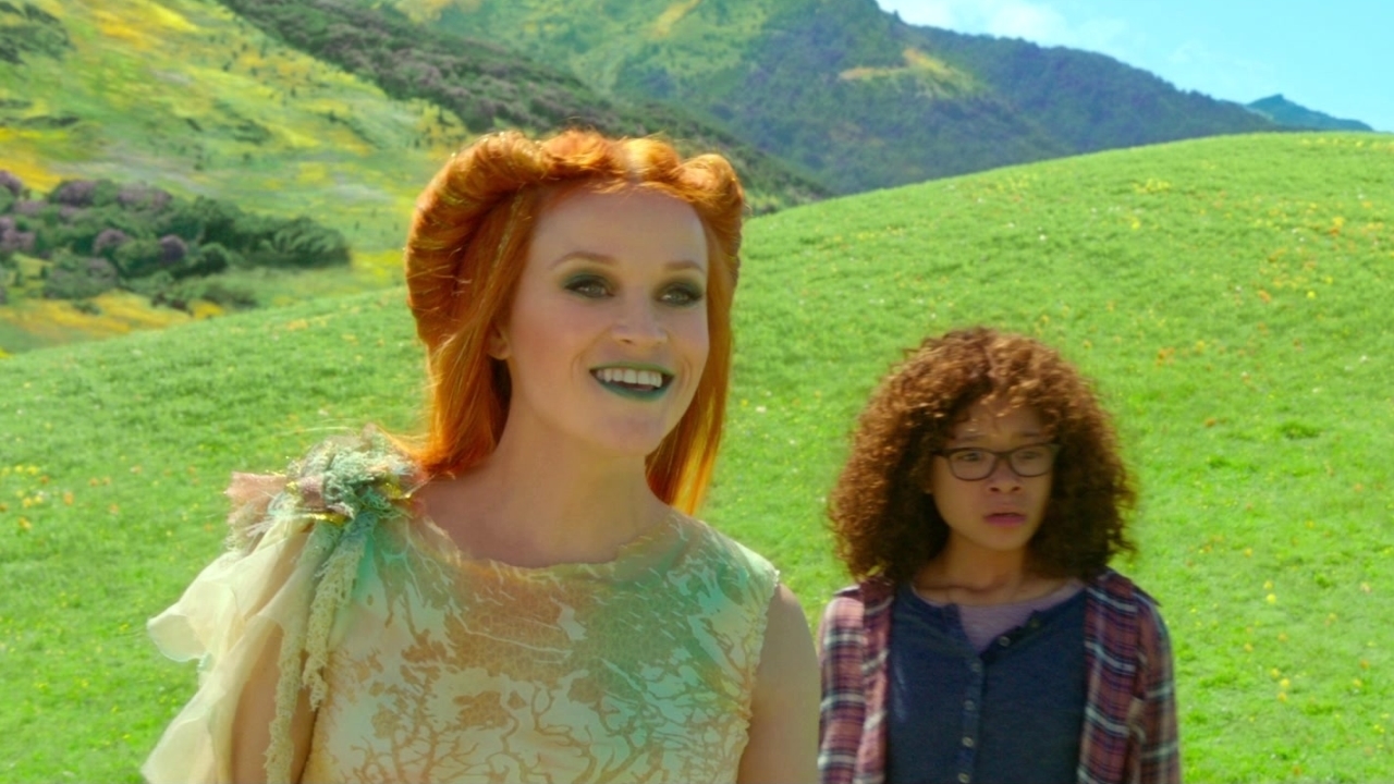Reese Witherspoon in a fantastical dress standing in a grassy meadow with Storm Reid in casual clothing in the background