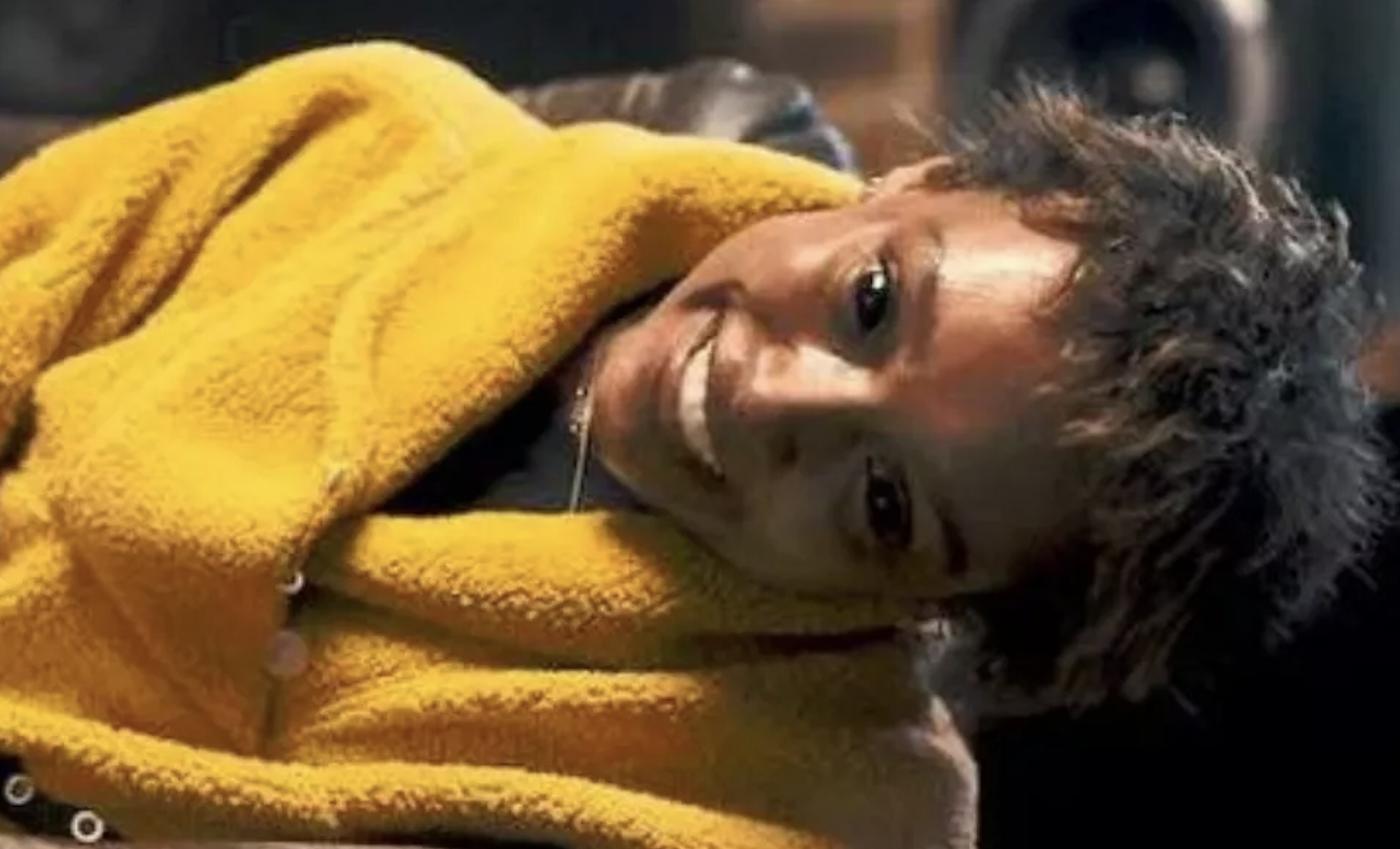 A smiling person wrapped in a fluffy blanket tilts their head playfully