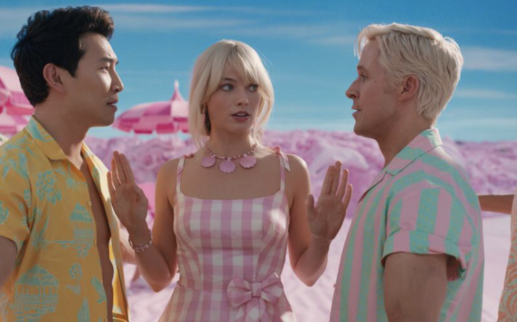 Simu Liu, Margot Robbie, and Ryan Gosling stand in a pink-themed outdoor setting. Margot wears a pink checkered dress with a matching necklace