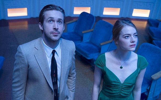 Ryan Gosling in a beige suit and tie stands next to Emma Stone in an elegant green dress in an empty theater, both looking upwards