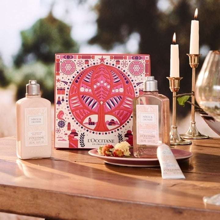 A display of L'Occitane en Provence products, including two bottles, a tube of cream, and a decorative box, set on a wooden table with candles and a wine glass