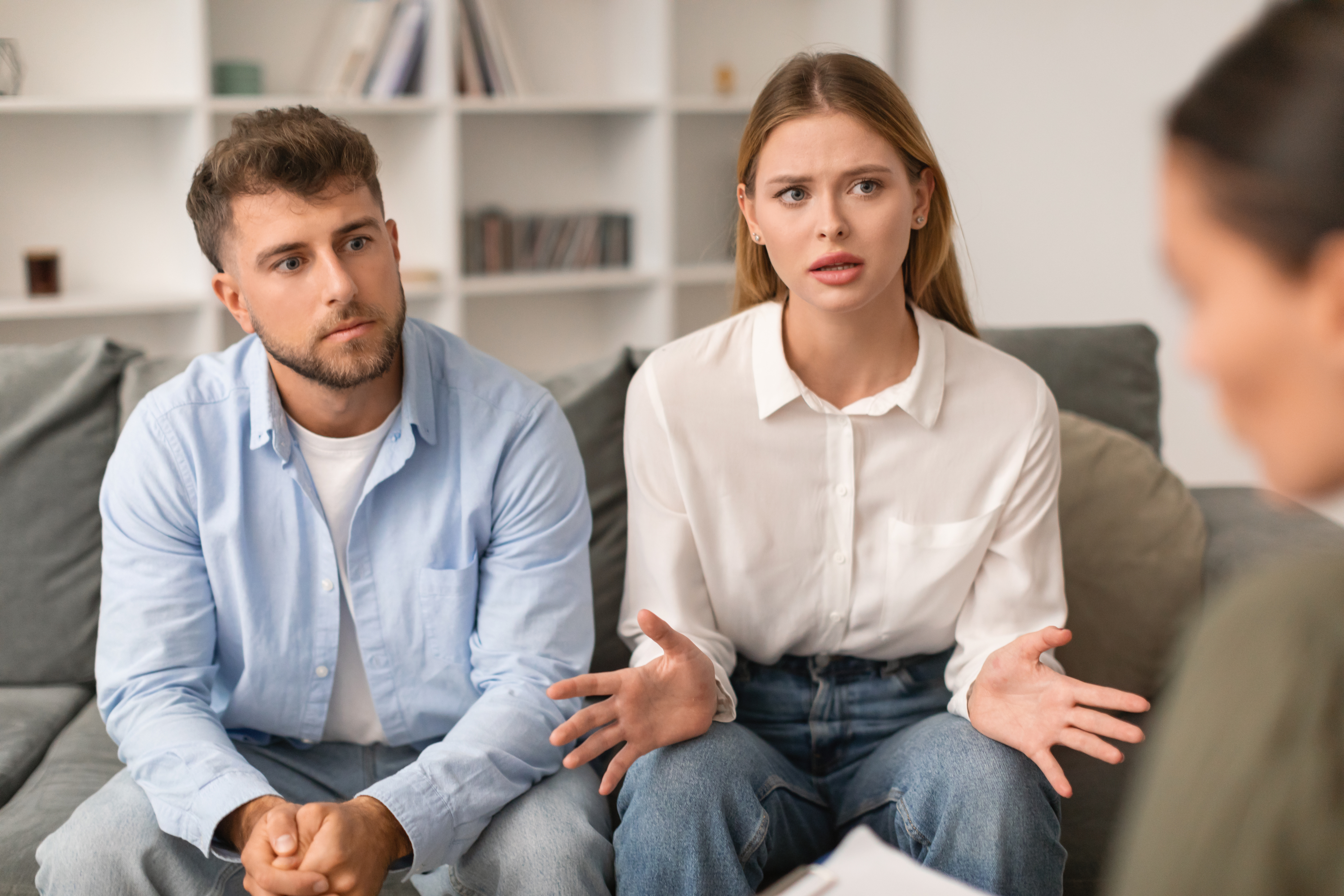 A man and a woman sit on a couch, engaged in a serious conversation with an unseen person. The woman gestures animatedly, while the man looks concerned