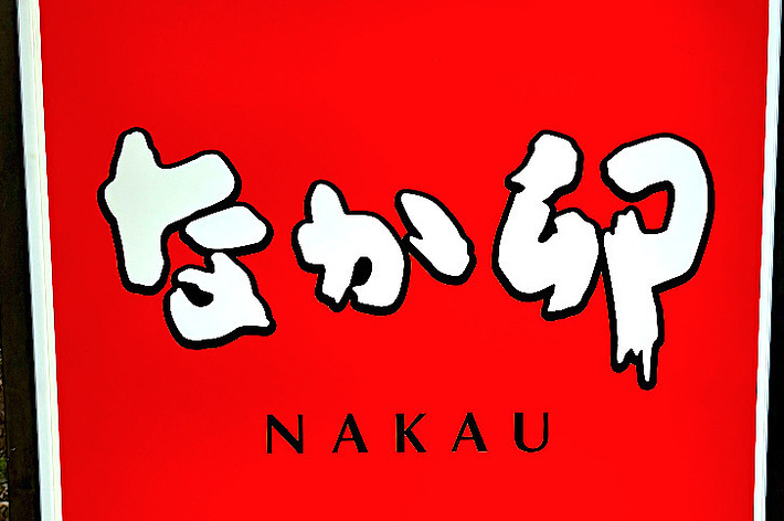 A street sign displays &ldquo;Nakau,&rdquo; advertising &ldquo;donburi&rdquo; (rice bowl dish) and &ldquo;Kyoto-style udon&rdquo; (noodle soup). The sign is near a sidewalk with buildings in the background