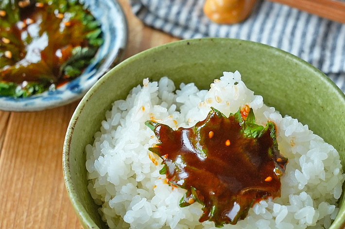 ご飯の上にごまとタレがかかった葉がのっている。背景には同じタレがかかった葉がさらに一枚、小皿にのせられている。