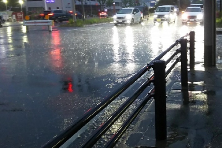 雨が降り、車道に車が走っている夜の街の風景。道路や歩道に水たまりができている。
