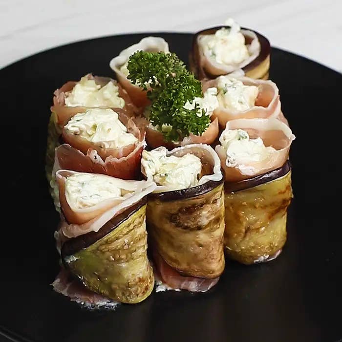 黒い皿の上に盛り付けられたクリームチーズとパセリを伴うナスと生ハムのロール。