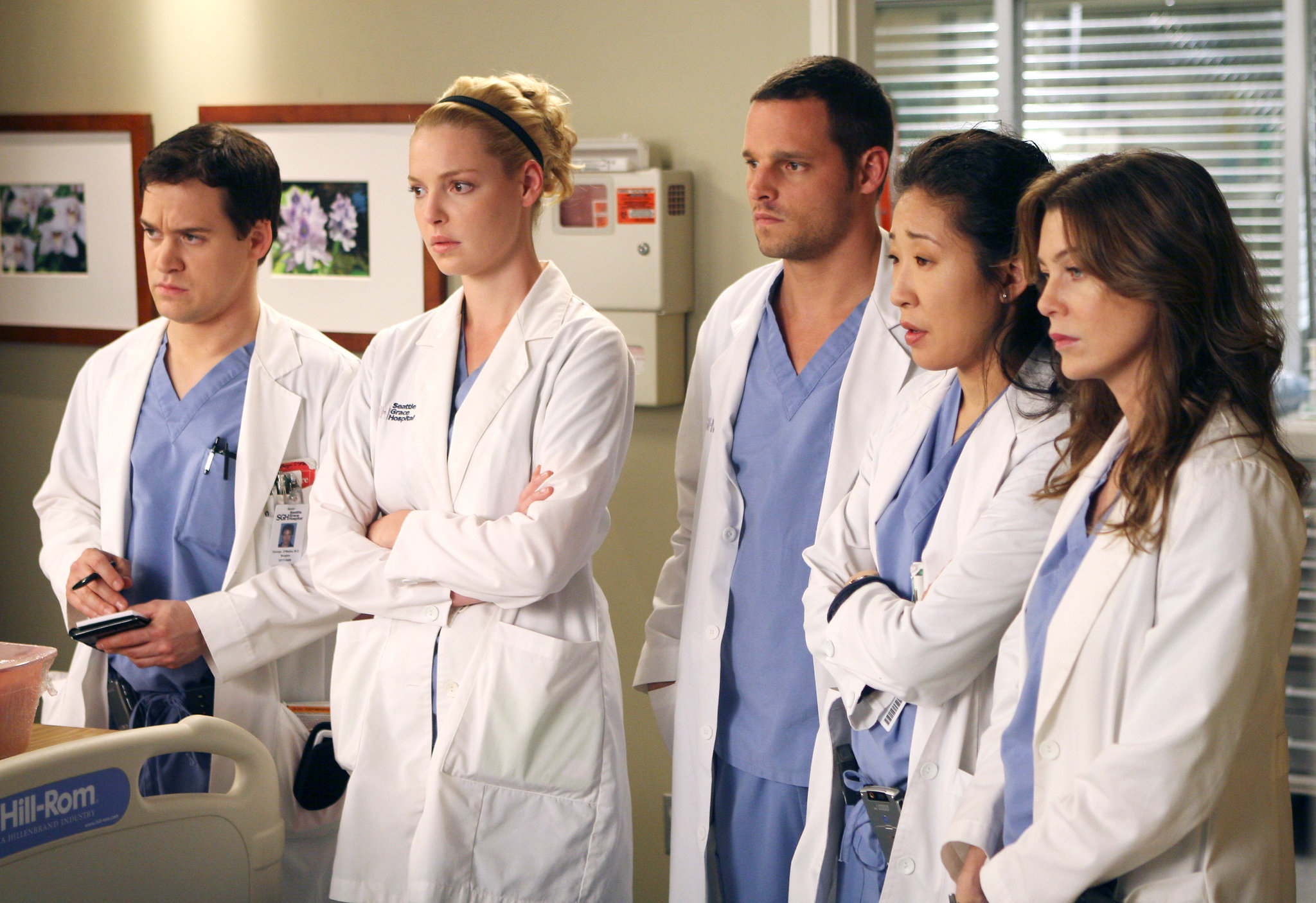 Patrick Dempsey, Katherine Heigl, Justin Chambers, Sandra Oh, and Ellen Pompeo in scrubs and lab coats, standing in a hospital room, seemingly in deep thought