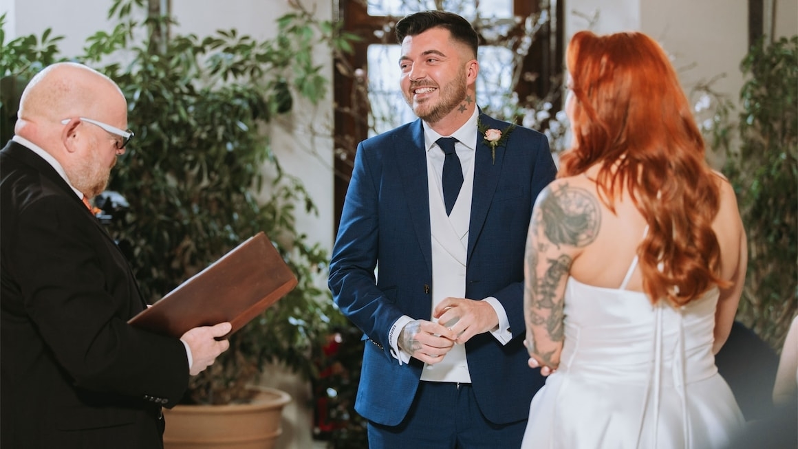 A wedding ceremony with a man in a blue suit, a woman in a white dress with tattoos, and an officiant holding a book