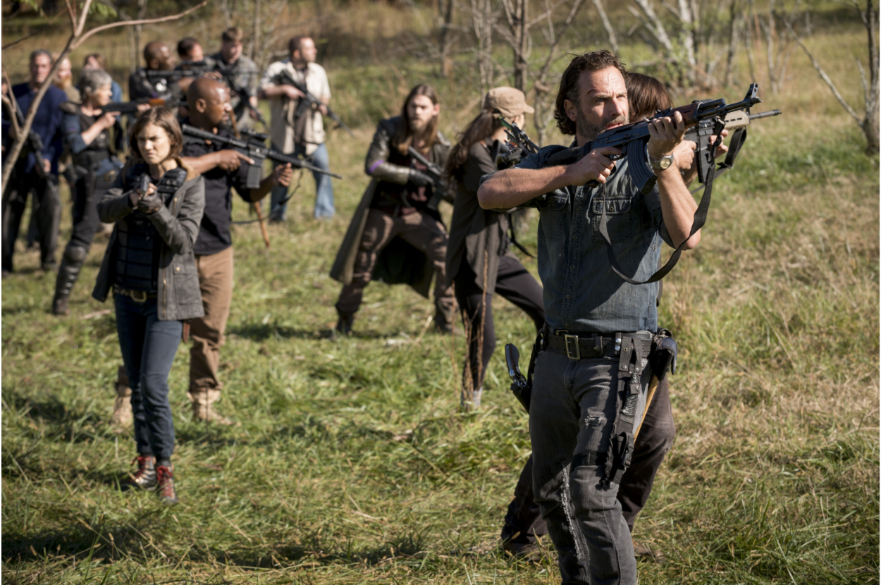 A group of people, including Andrew Lincoln, Lauren Cohan, and Norman Reedus, stand in a field holding weapons, appearing ready for a confrontation