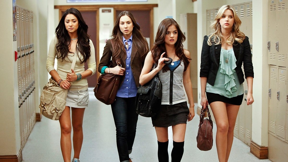 Shay Mitchell, Troian Bellisario, Lucy Hale, and Ashley Benson walk in a school hallway. They are casually dressed with handbags over their shoulders