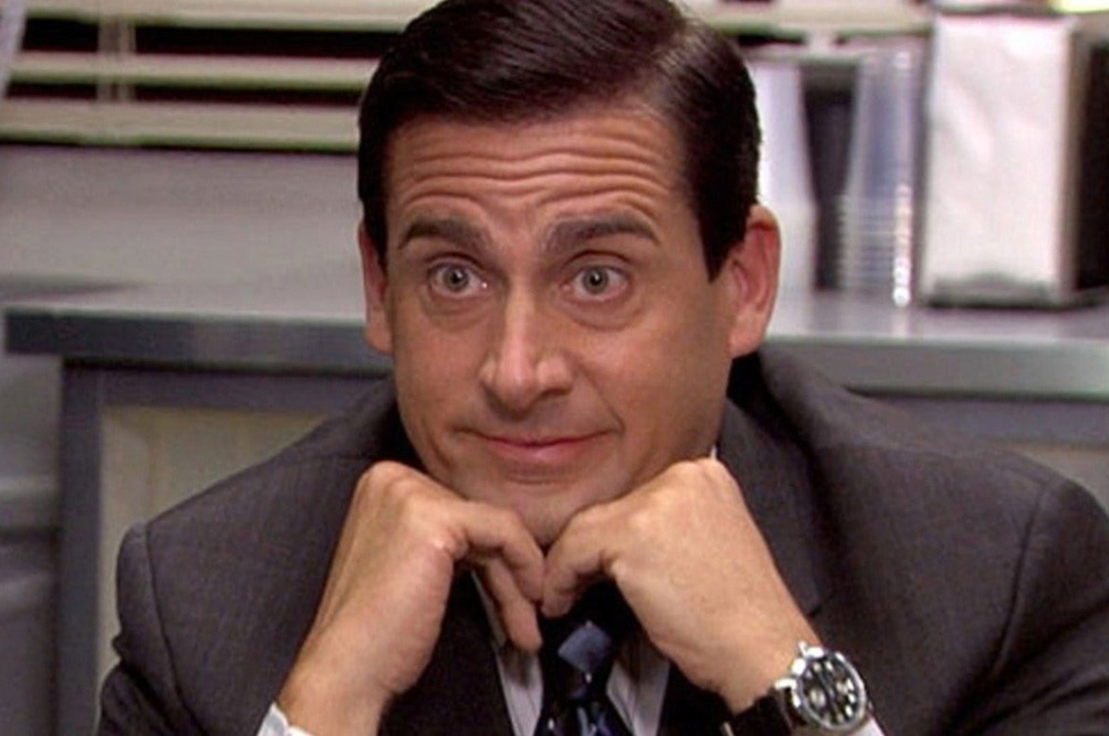 A man in a suit with his hands under his chin exhibits a thoughtful yet amused expression