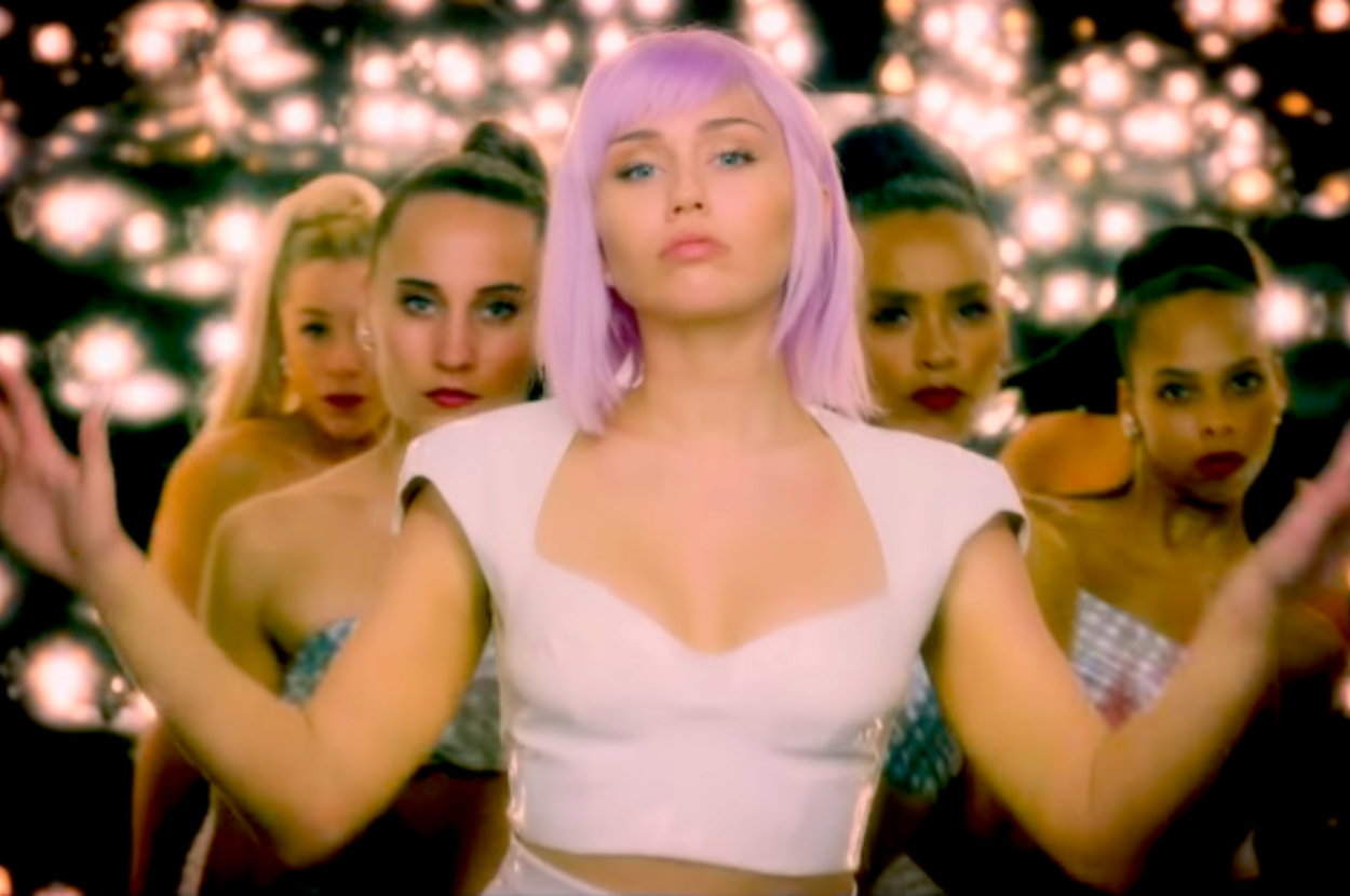 Miley Cyrus in a futuristic white outfit with a purple wig stands in front of four backup dancers with lights in the background