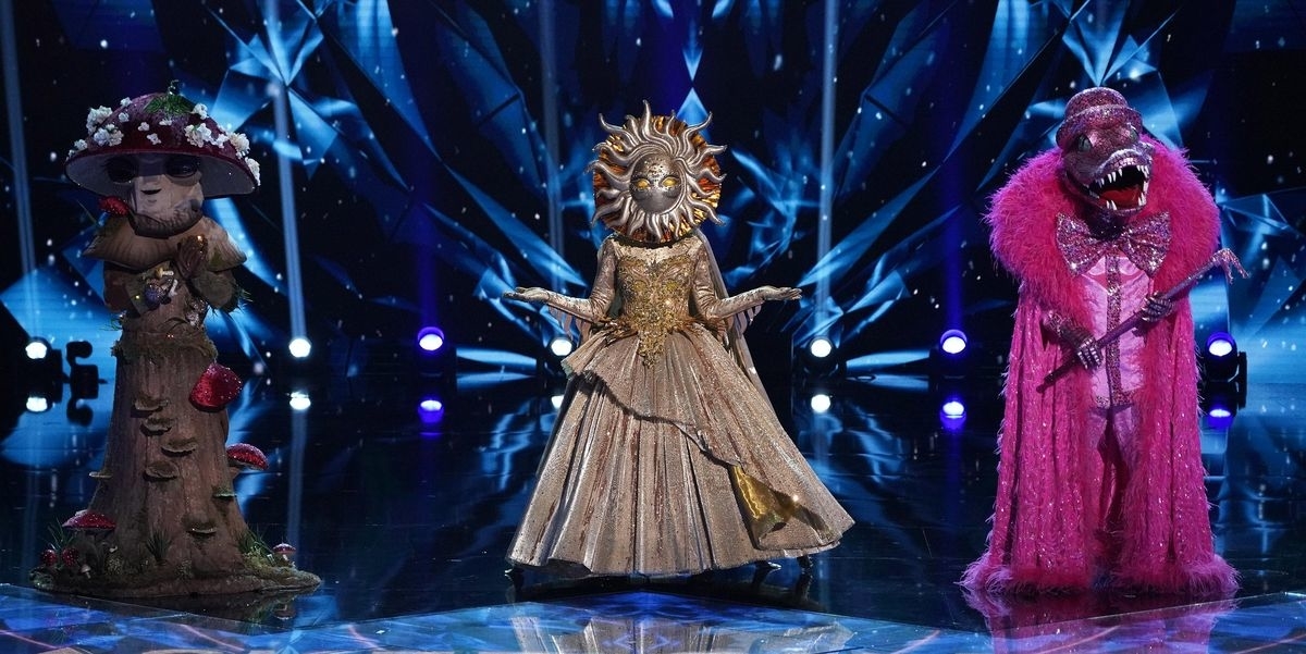 Three costumed characters on stage: Tree with mushroom accents, Sun in golden dress, and Pink Monster in a furry pink outfit with a pink feathered collar