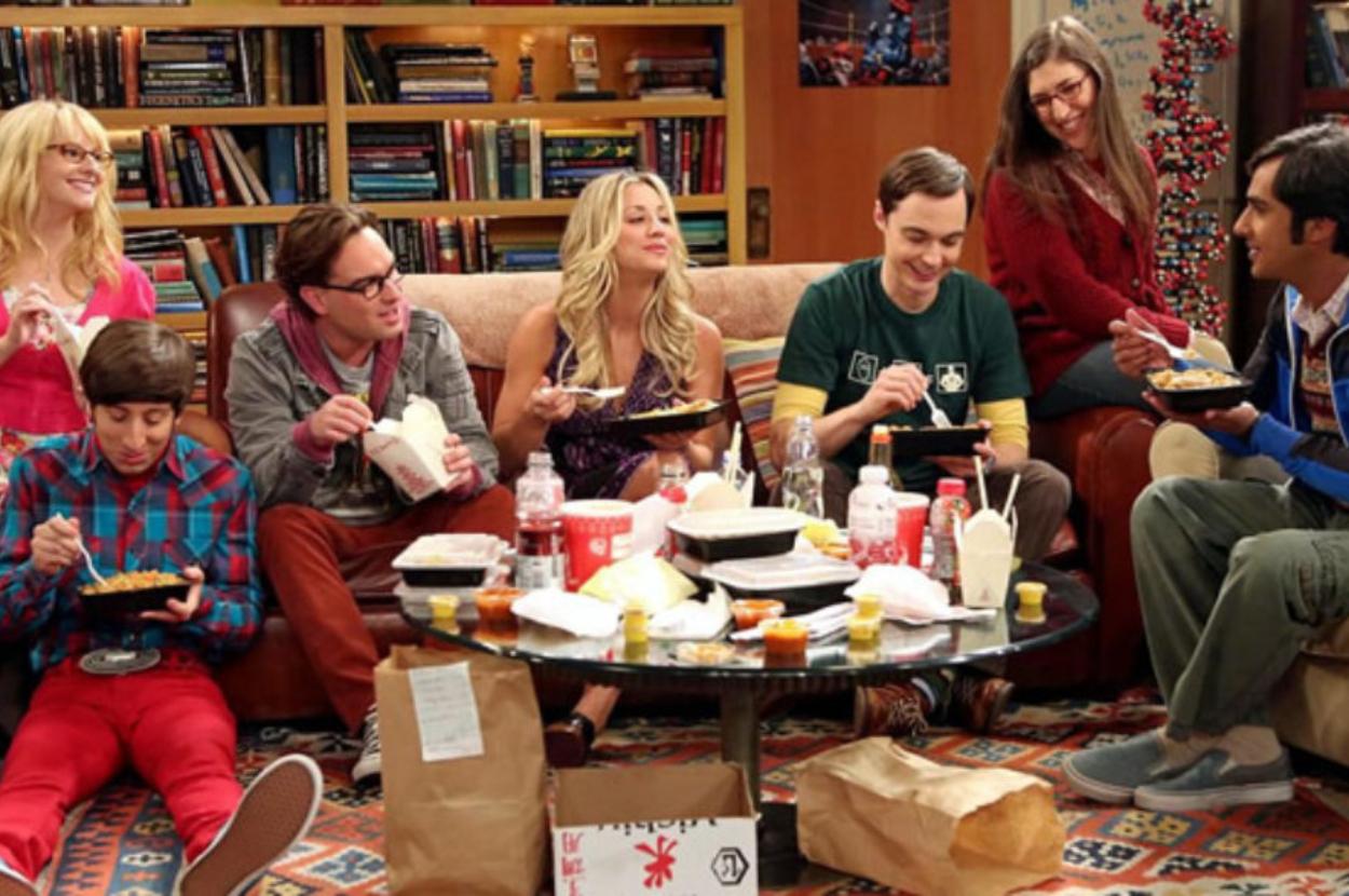 Melissa Rauch, Simon Helberg, Johnny Galecki, Kaley Cuoco, Jim Parsons, Mayim Bialik, and Kunal Nayyar sit in a living room eating takeout food, smiling, and laughing