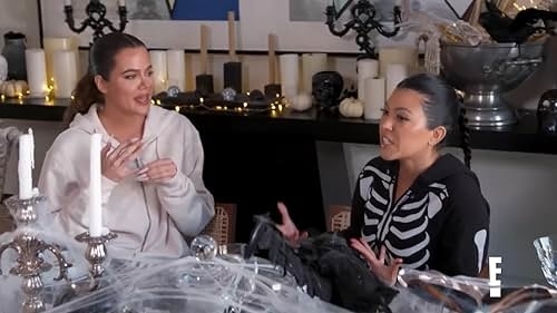 Khloé Kardashian and Kourtney Kardashian are talking while sitting at a table decorated with Halloween-themed items, including candles and skulls