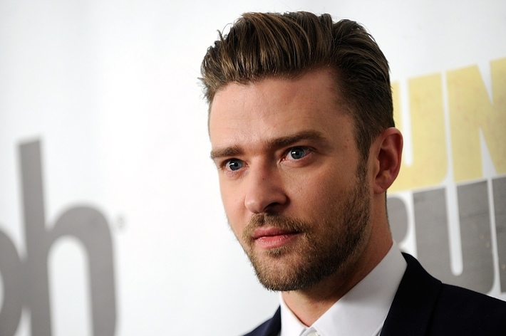 Justin Timberlake in a suit at a media event with a backdrop displaying partially visible text