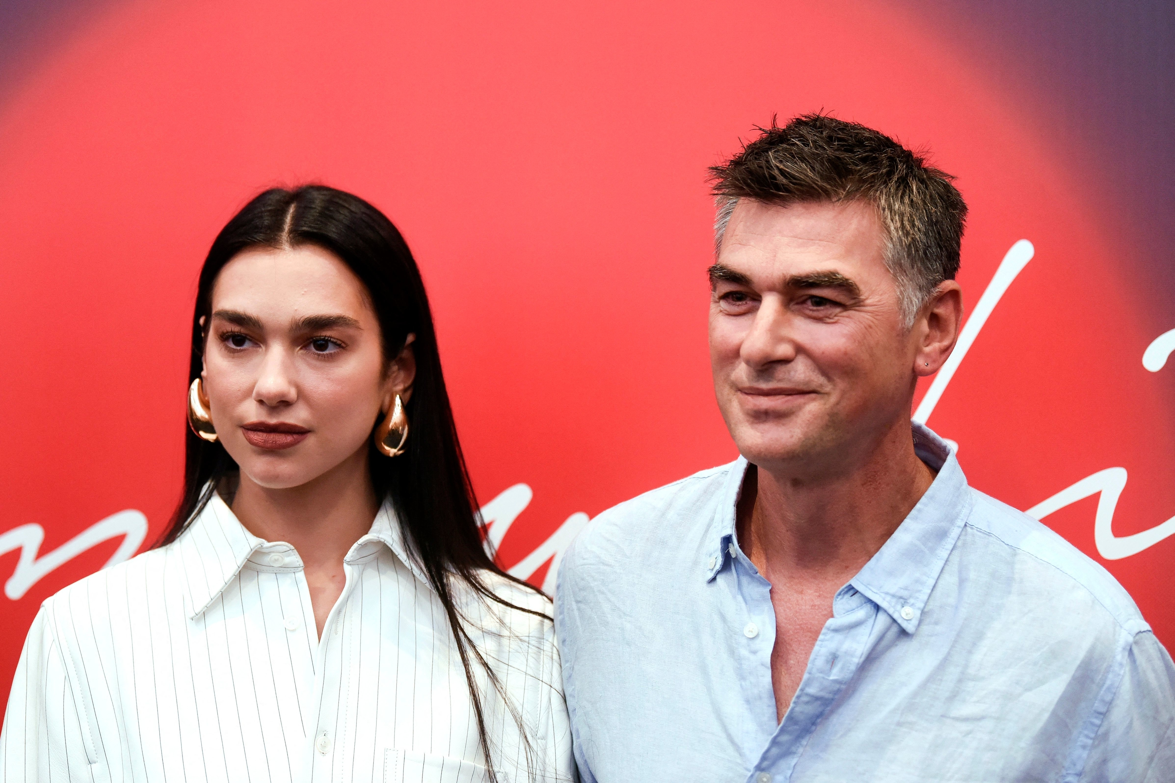 Dua Lipa in a striped blouse and large earrings, stands alongside her dad in a light shirt at a red-carpet event