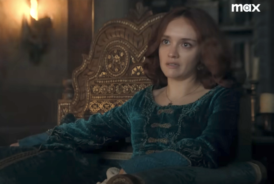 Olivia Cooke sits on an intricately designed throne-like chair, wearing a medieval-style dress