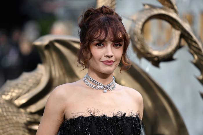 Olivia in a strapless, feathered dark dress and pearl choker necklace stands in front of a dragon-themed background at a public event