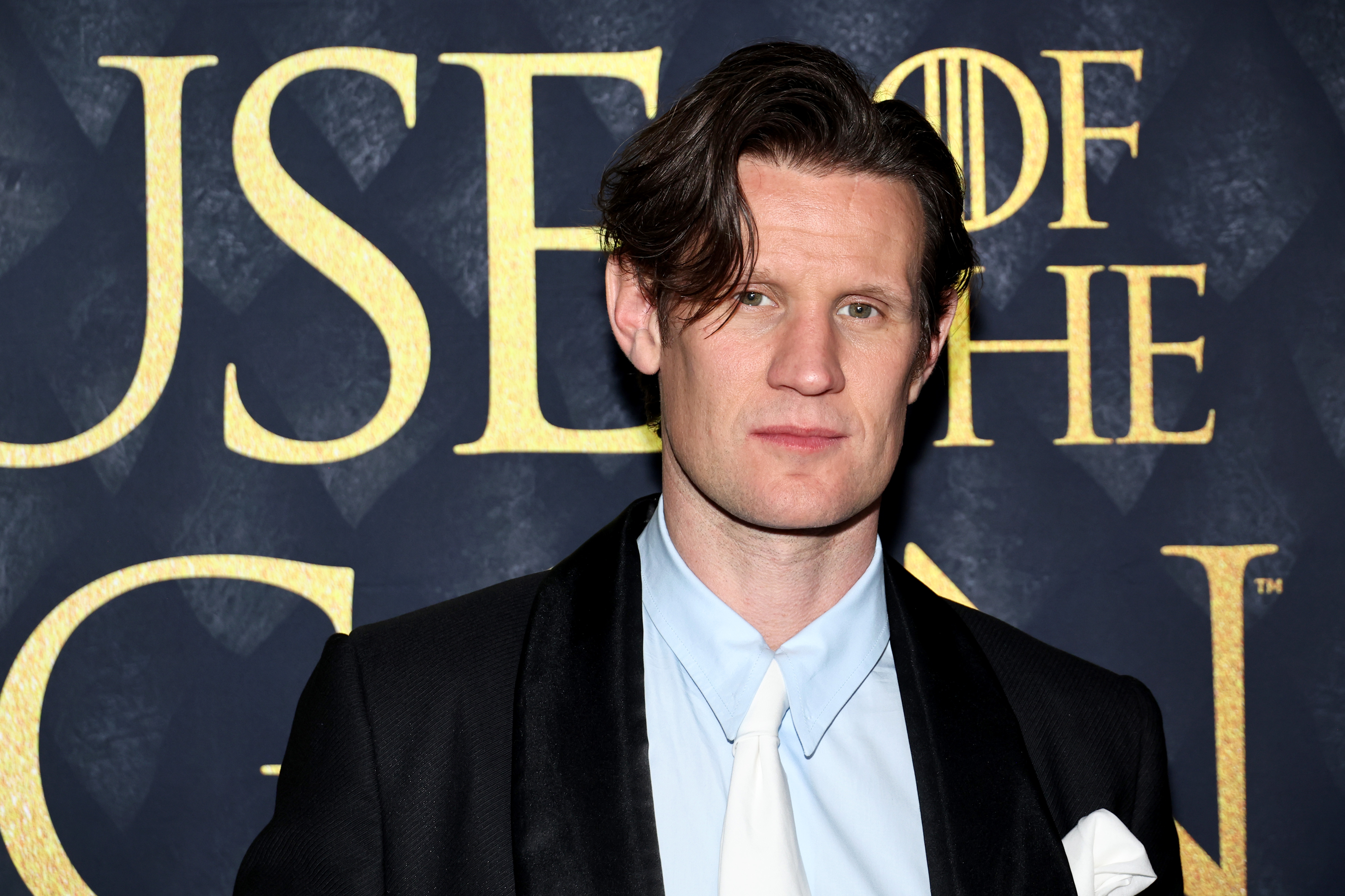 Matt Smith poses in front of a &quot;House of the Dragon&quot; promotional background, wearing a black blazer, white shirt, and white tie