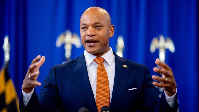 Wes Moore delivers a speech, wearing a navy suit, white shirt, and orange tie, with a blue backdrop featuring flags