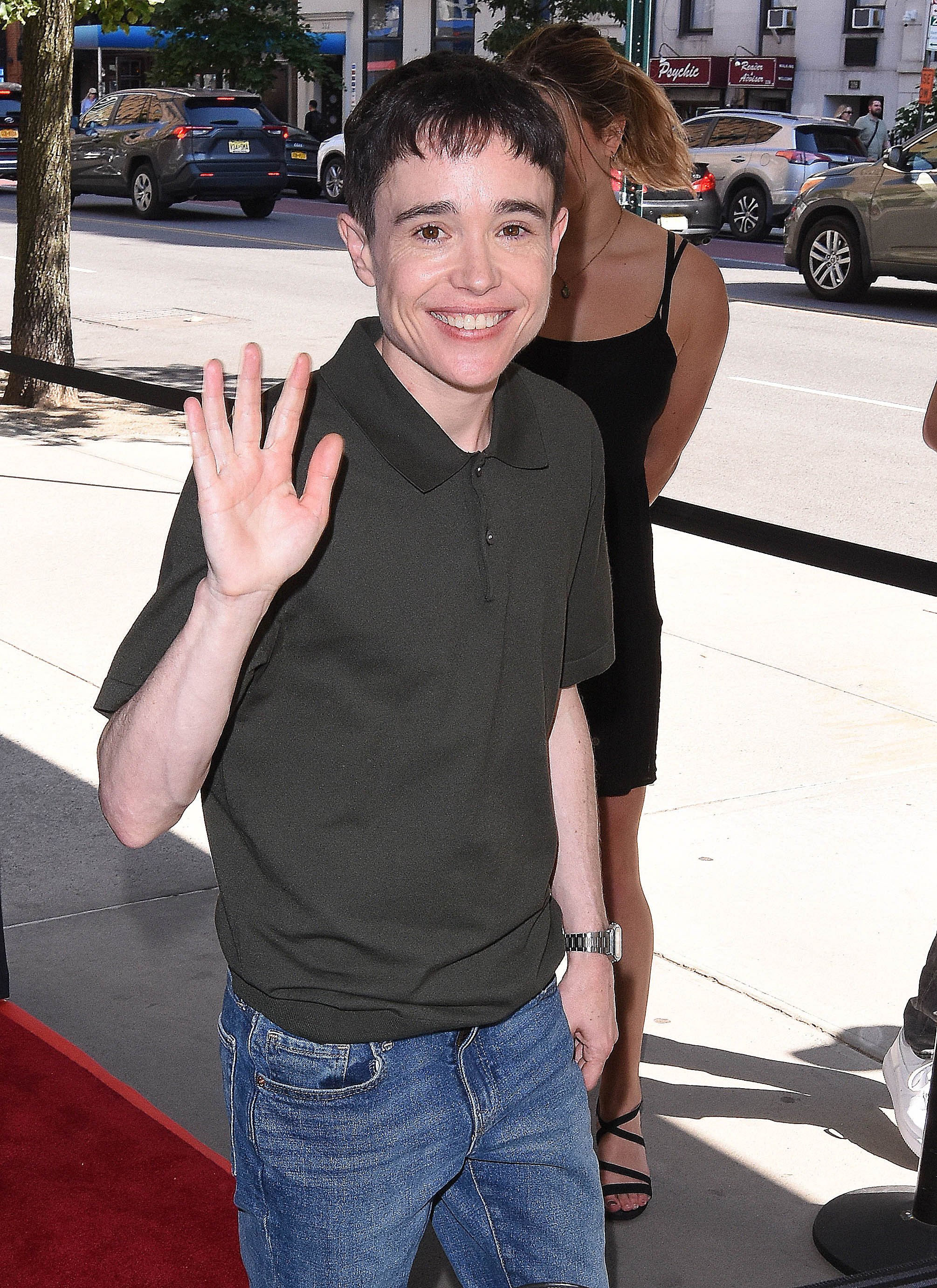 Elliot Page waves and smiles on a sidewalk near a red carpet. He is wearing a casual dark polo shirt and jeans