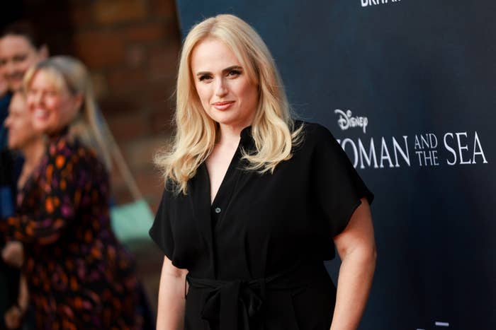 Rebel Wilson stands on the red carpet at a Disney event, wearing a stylish black dress. Other attendees are in the background. Text on the backdrop says &quot;WOMAN AND THE SEA&quot;