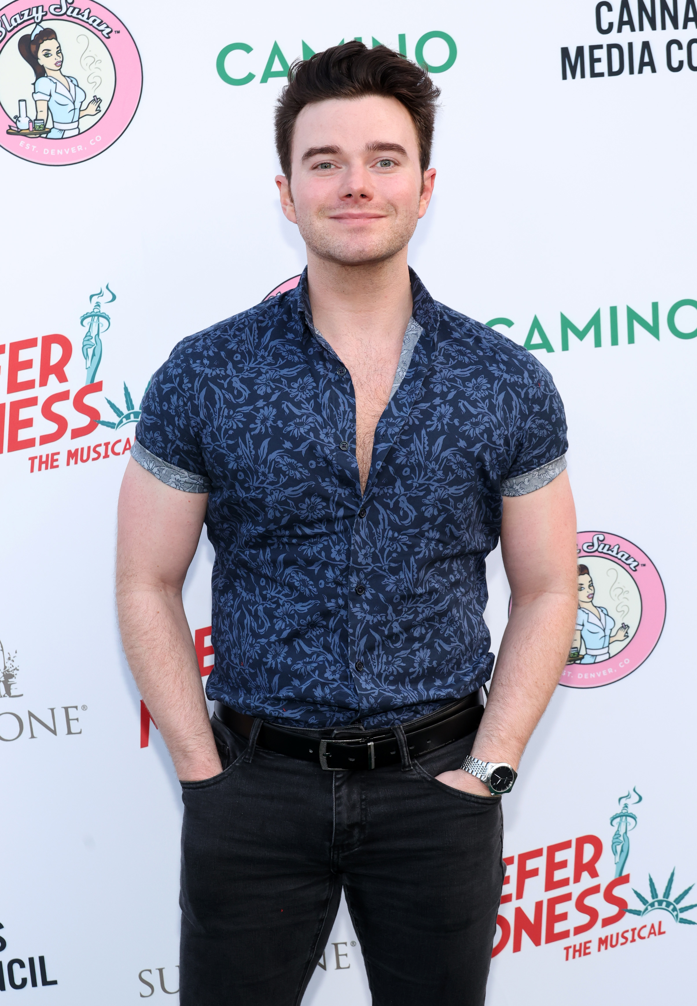 Chris Colfer stands on the red carpet wearing a patterned short-sleeved shirt, dark jeans, and a belt at an event backdrop for Reefer Madness: The Musical
