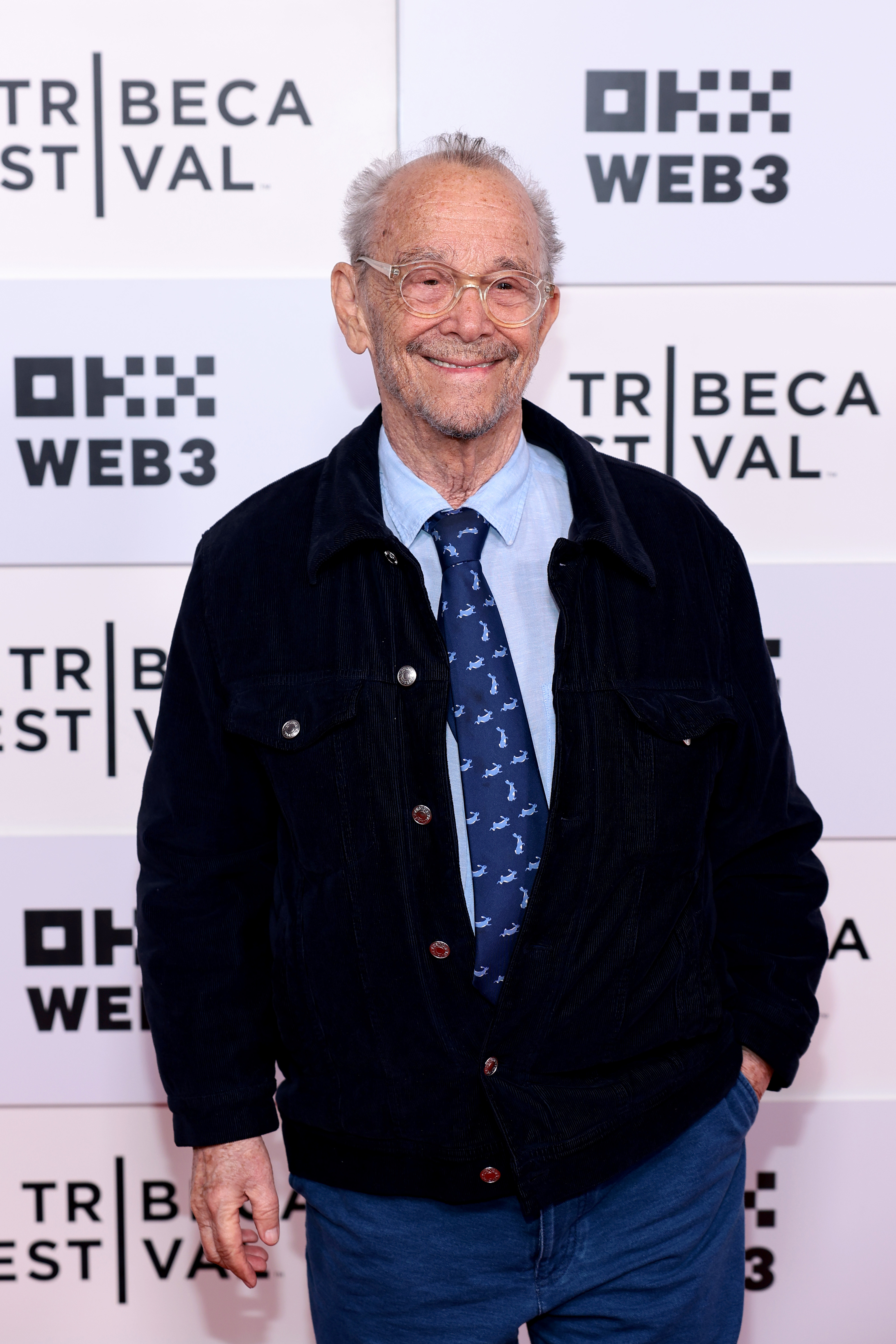 An older man in a casual outfit with glasses and a patterned tie, standing on the red carpet at the Tribeca Festival