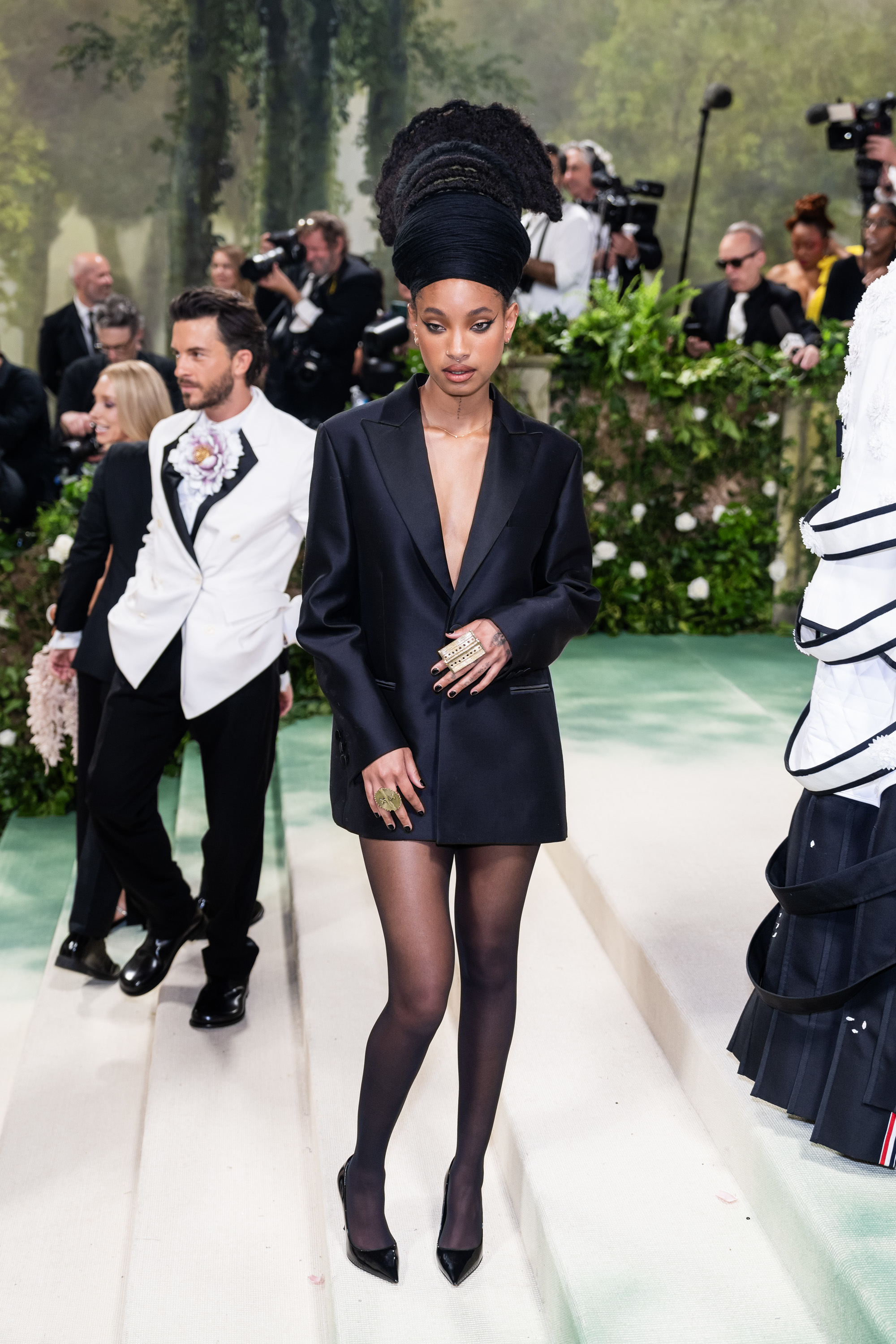 Two people on a red carpet. Willow Smith wears a sleek, black oversized blazer, black tights, and black heels. Another person in a white tuxedo jacket is in the background