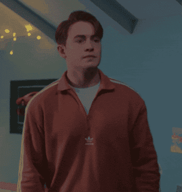 Kit Connor, wearing a collared, long-sleeve shirt with stripes on the sleeves, stands indoors, looking to the side with a thoughtful expression