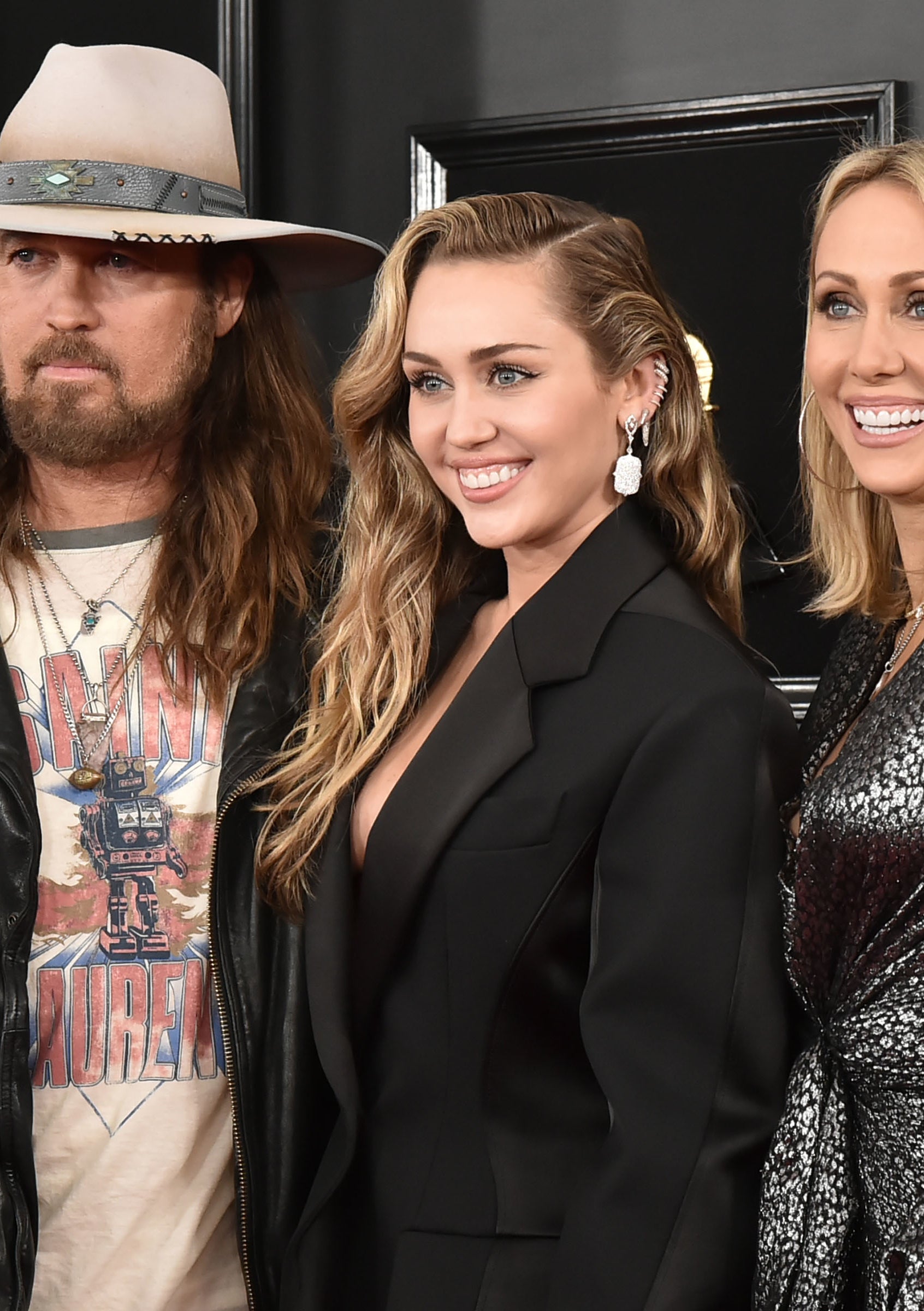 Billy Ray Cyrus, Miley Cyrus, and Tish Cyrus pose together; Billy Ray wears a hat and leather jacket, Miley is in a black blazer, and Tish wears a metallic dress