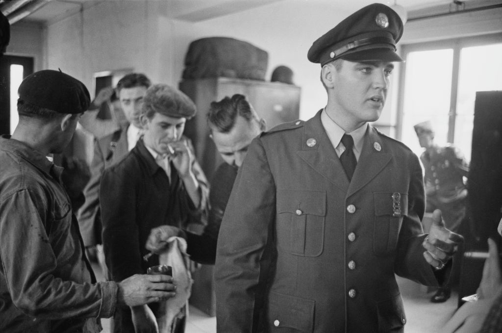 Elvis Presley in military uniform, stands inside a room with several men in casual clothes in the background