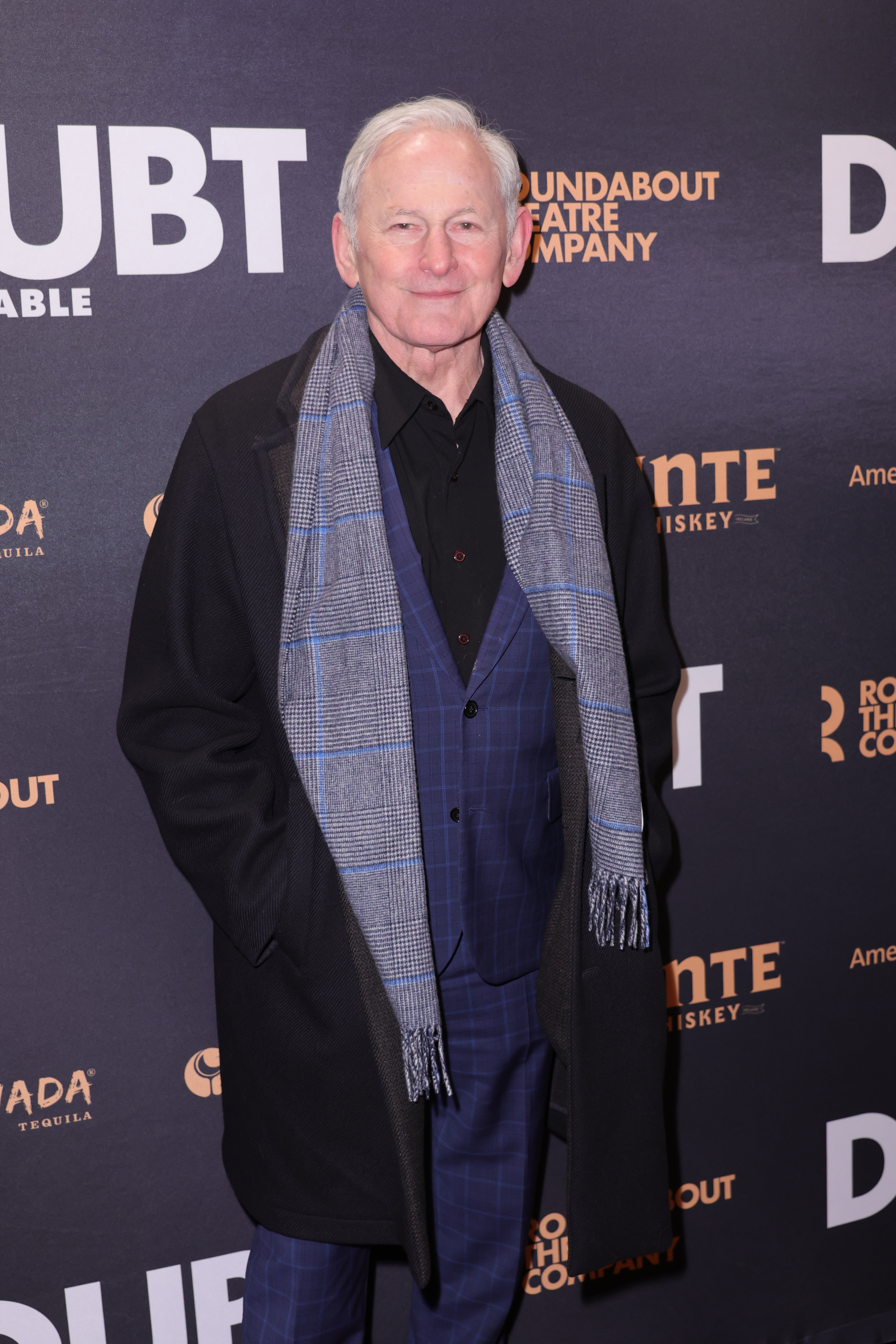 Victor Garber smiles while wearing a dark coat, a gray plaid scarf, and a navy suit at a Roundabout Theatre Company event