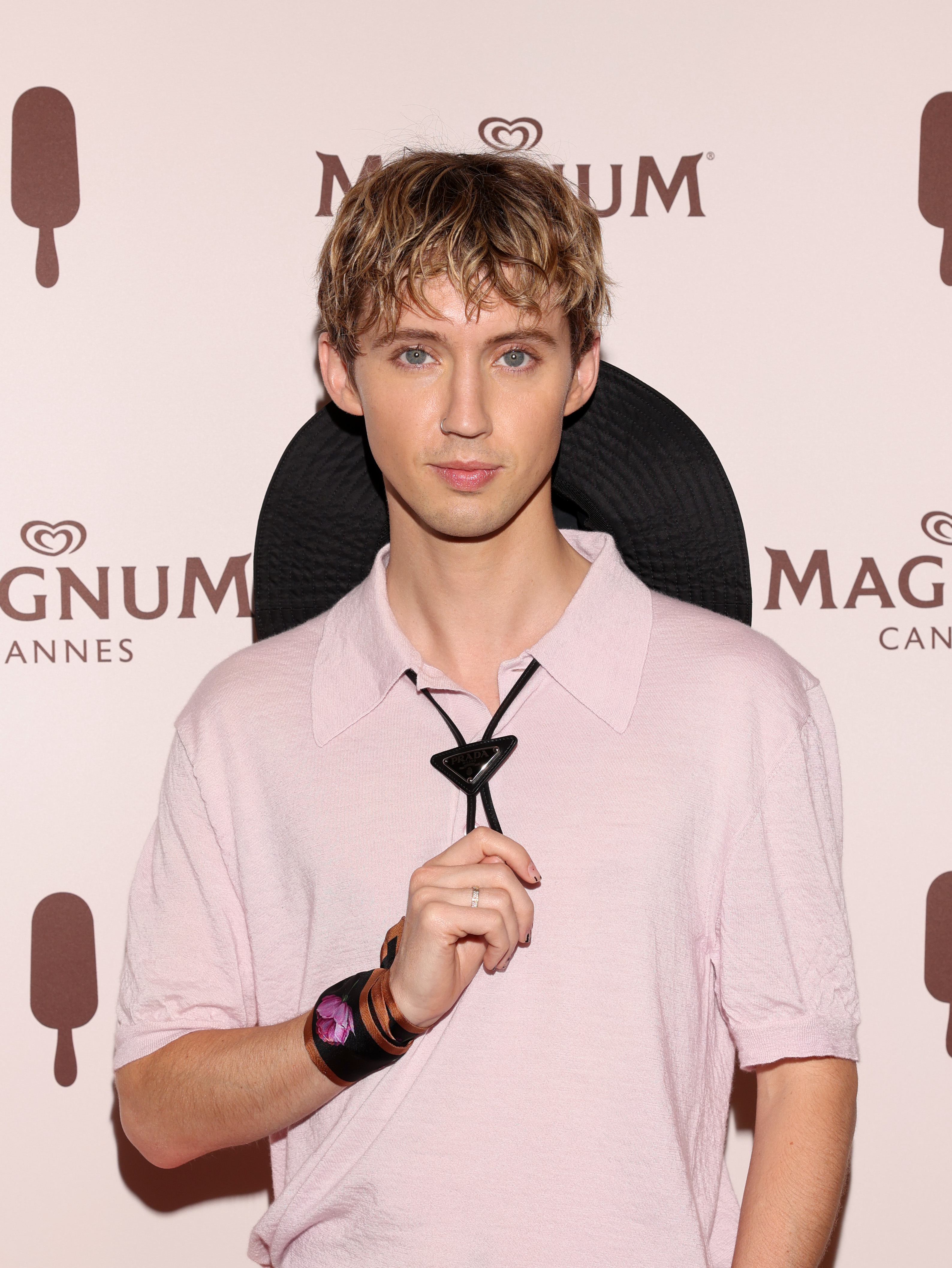 Troye Sivan at a Magnum Cannes event, wearing a short-sleeve shirt with a large black floppy hat resting behind his head, holding the hat's chin strap
