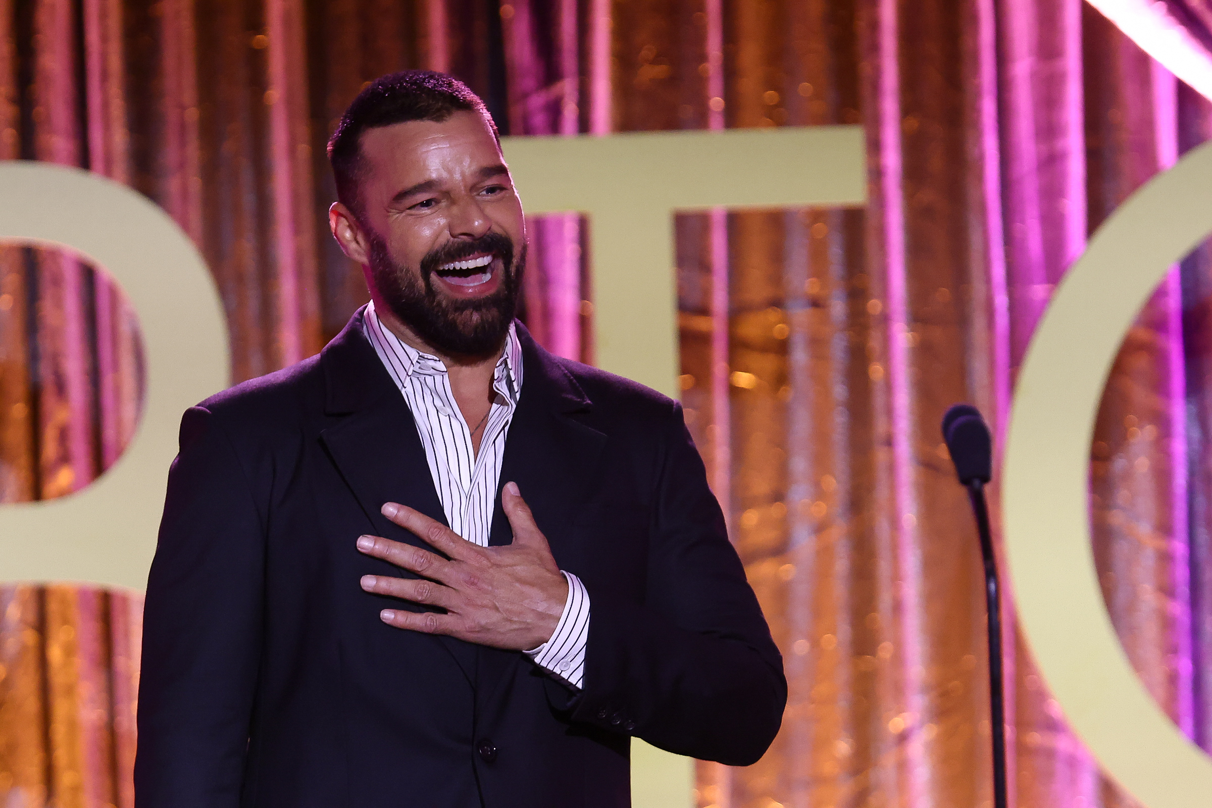Ricky Martin stands on stage, smiling and holding his hand to his chest, wearing a dark suit and striped shirt. Background: large letters and a curtain