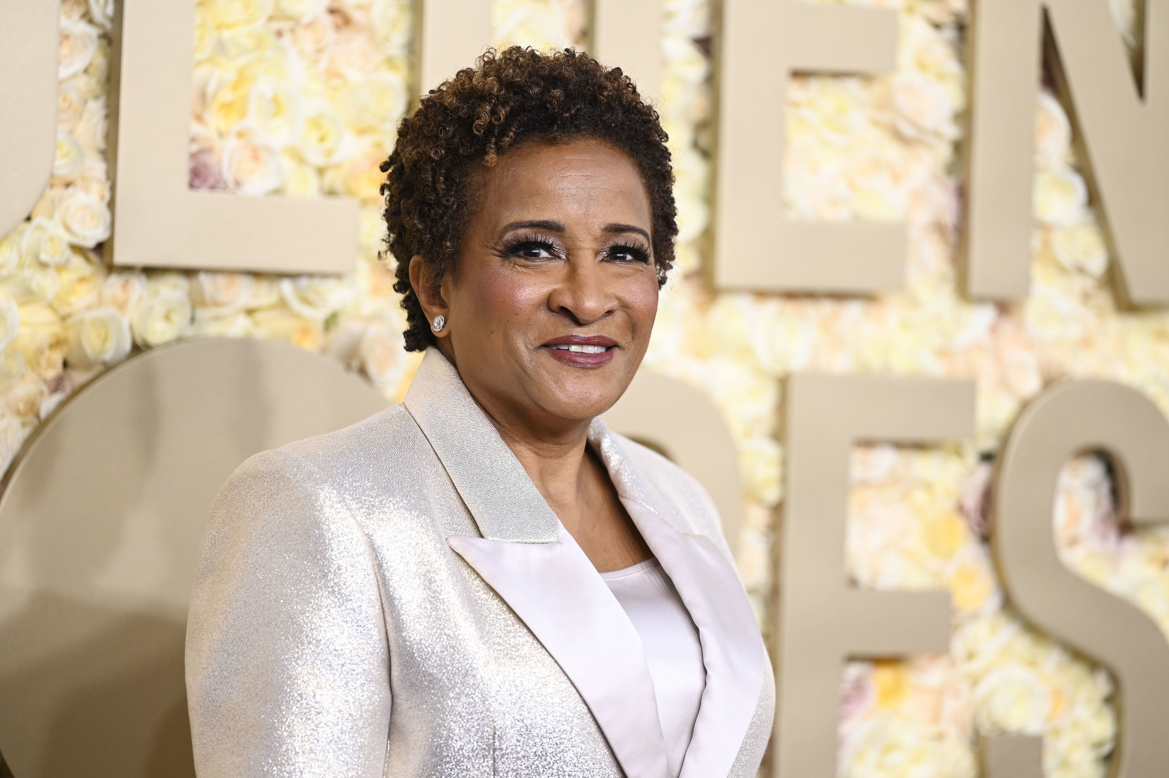 Wanda Sykes smiles while wearing a light-colored jacket and blouse, standing in front of a flower-covered background with large letters