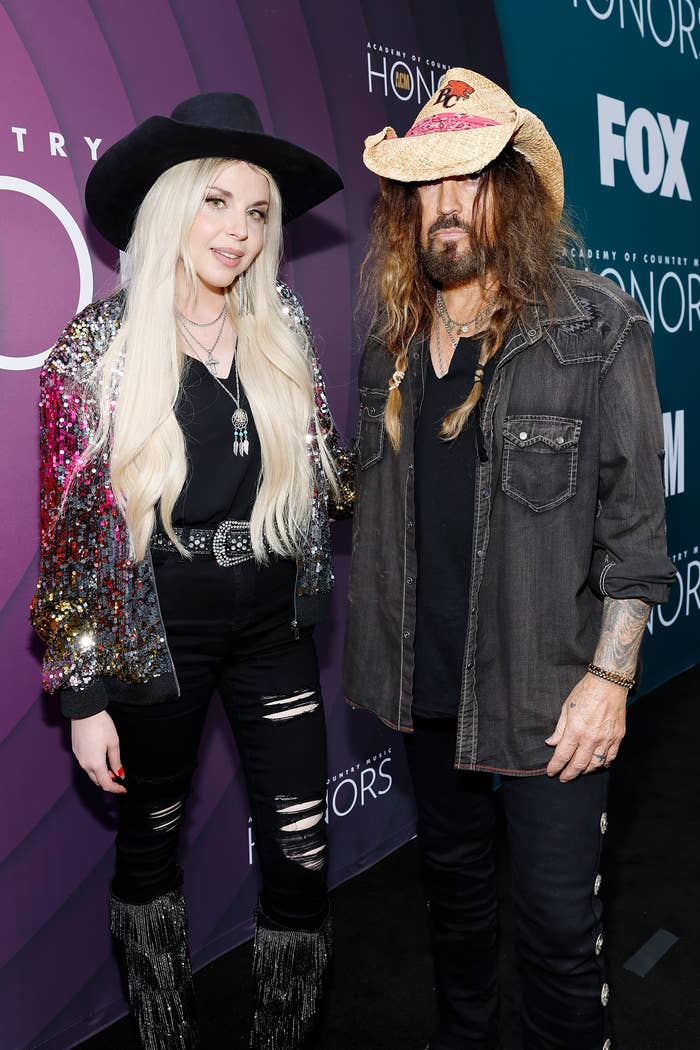 Firerose and Billy Ray Cyrus at an event, standing side by side. Kesha wears a sequined jacket and black outfit; Billy Ray Cyrus wears a cowboy hat, denim jacket, and black shirt