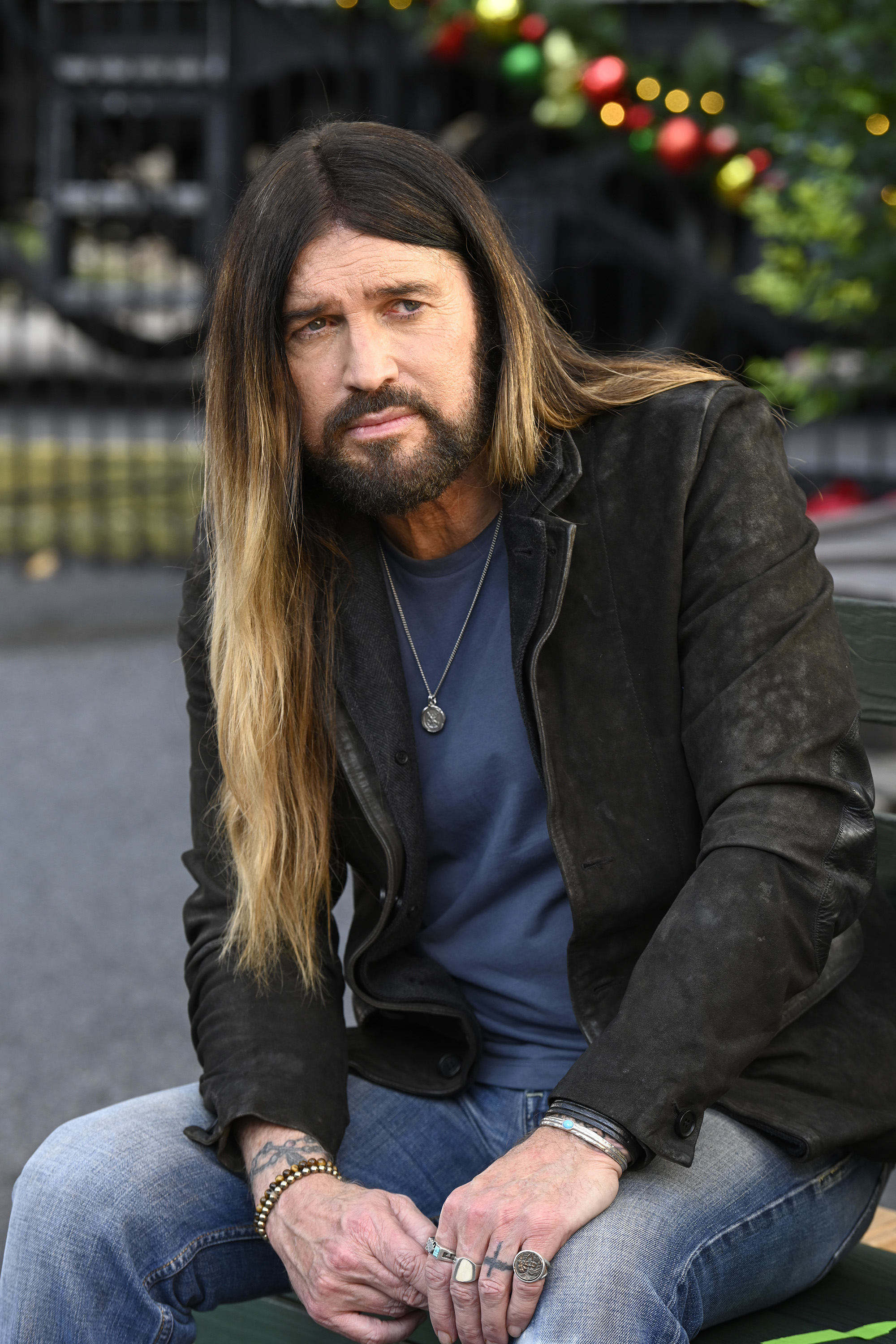 Billy Ray Cyrus is sitting outdoors, wearing a black leather jacket, a blue shirt, and jeans. He has long hair and multiple rings on his fingers