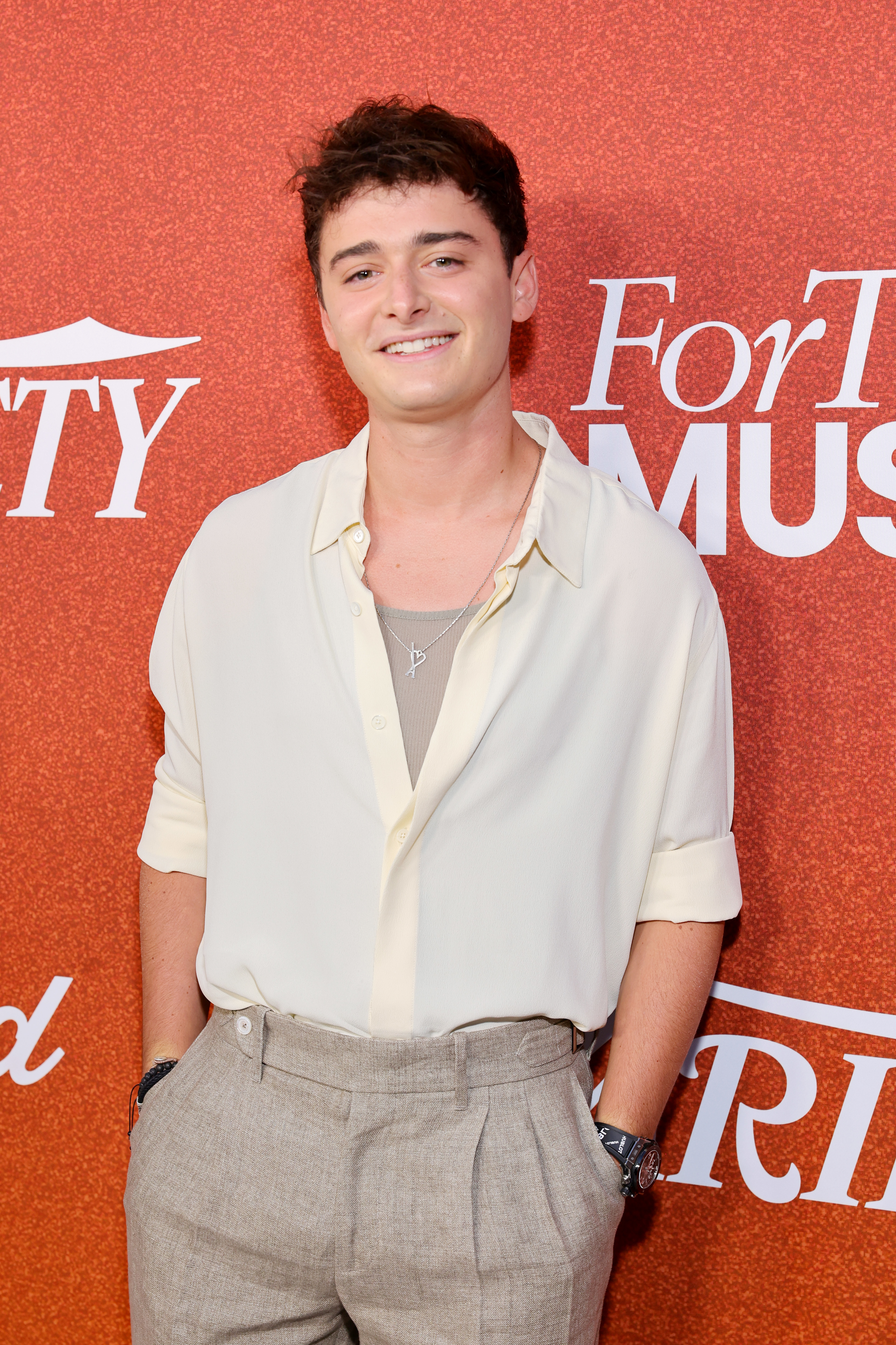 Noah Schnapp smiling on a red carpet, wearing a loose white shirt over a gray tank top, and gray pants