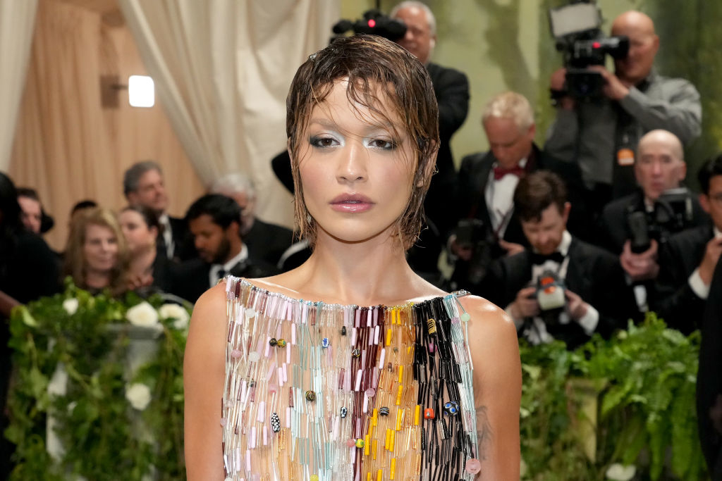 Woman with short, wet hair in a colorful, beaded dress attends a formal event with photographers and attendees in the background