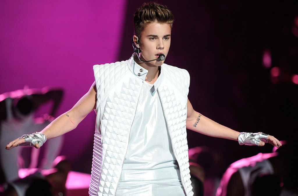 Justin Bieber performs on stage wearing a white quilted vest and white pants, holding a microphone near his mouth