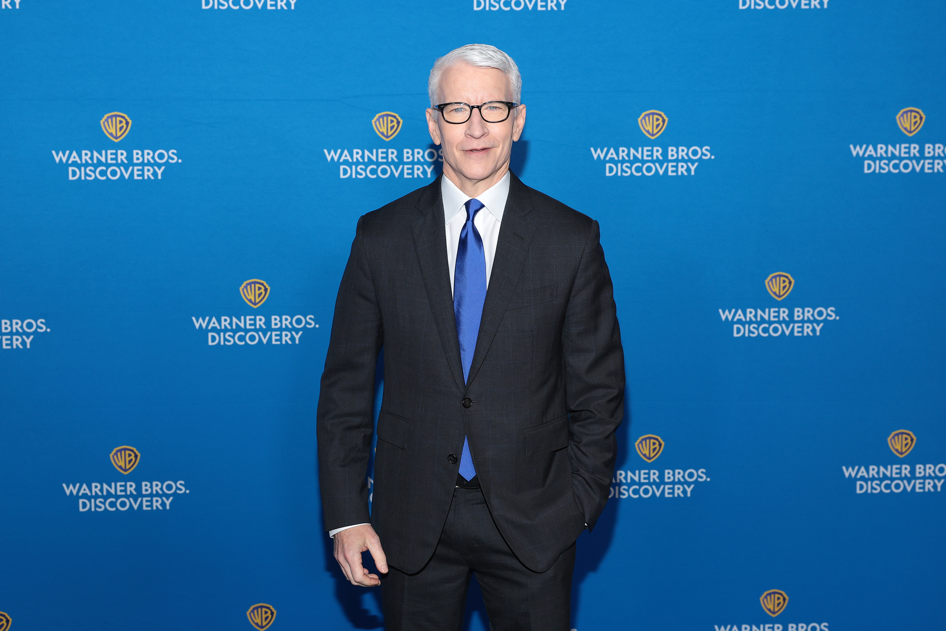 Anderson Cooper in a suit and tie poses in front of a Warner Bros. Discovery backdrop
