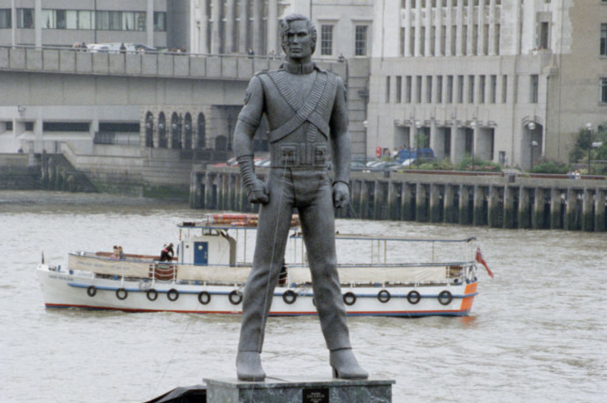 Statue of Michael Jackson in a military-style outfit with hands on hips, overlooking a river with a boat and buildings in the background