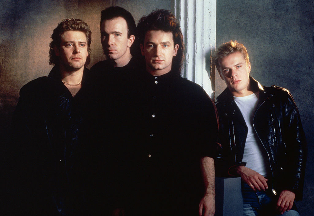 A group photo of U2 members: Adam Clayton, Larry Mullen Jr., Bono, and The Edge, all dressed in casual black and white attire against a neutral background