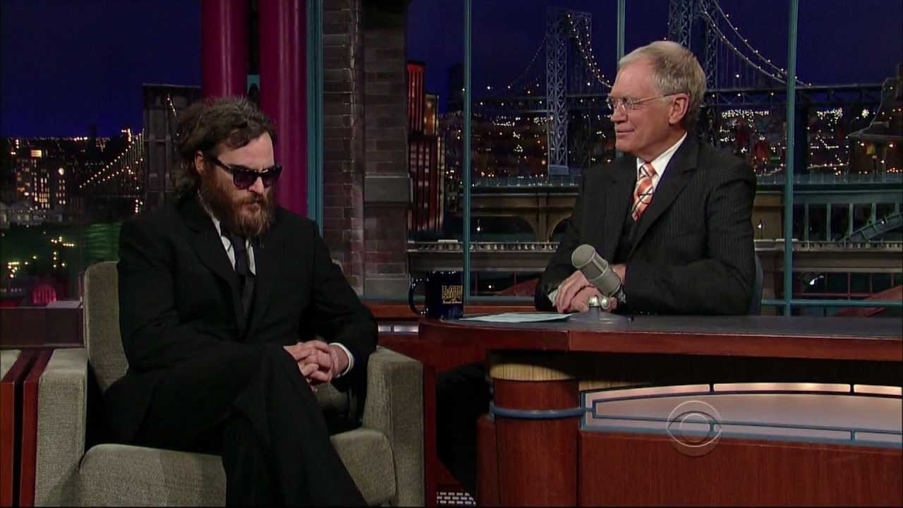 Joaquin Phoenix, wearing a dark suit and sunglasses, sits on a talk show set, speaking with host David Letterman, who is dressed in a suit and tie