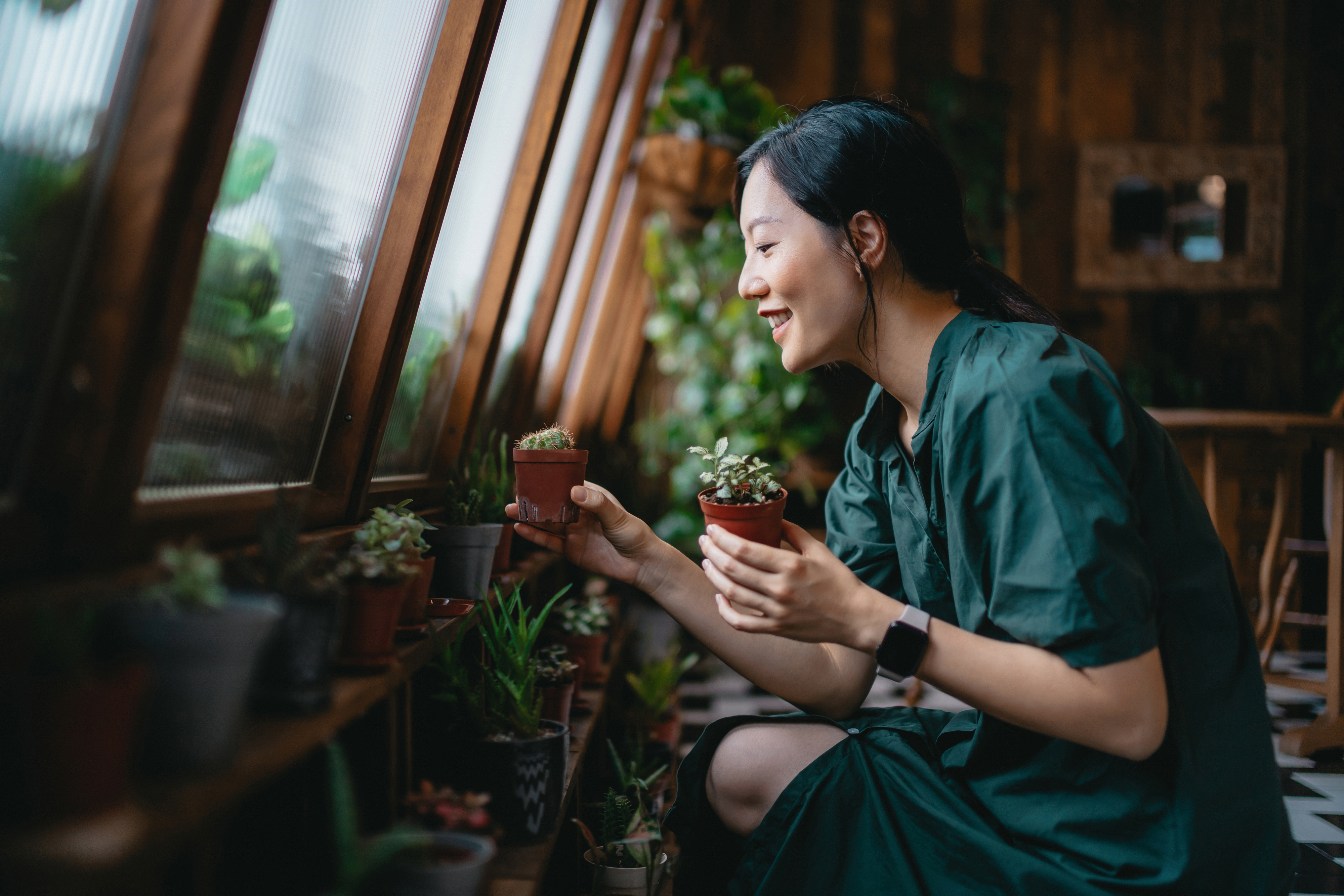A woman in a green dress, holding small potted plants, smiles as she looks out a window surrounded by greenery